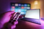Hand mit Fernbedienung vor einem Smart-TV und Tablet im Wohnzimmer