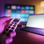 Hand mit Fernbedienung vor einem Smart-TV und Tablet im Wohnzimmer
