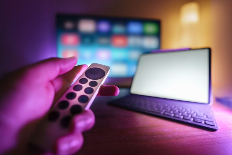 Hand mit Fernbedienung vor einem Smart-TV und Tablet im Wohnzimmer